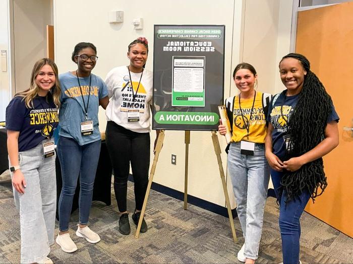 Five female students stand around a tripod holding a conference poster.